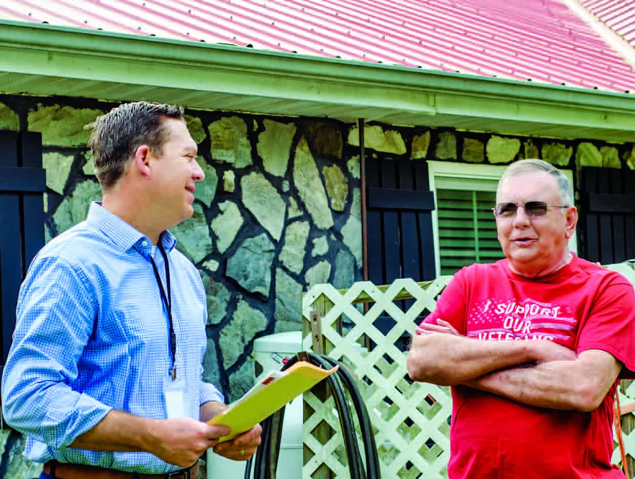 Two people converse outside a stone house with a red roof. One holds a file folder, the other wears a red shirt.