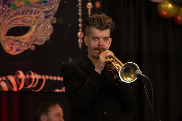 A person plays the trumpet on stage with a masquerade-themed backdrop, near a microphone, alongside another person partially visible in the background.