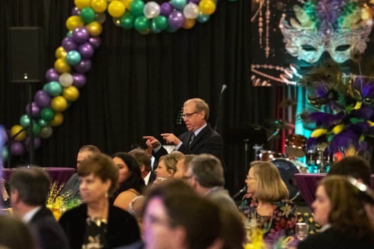 A festive gathering with colorful balloons, masks, and a person speaking to seated guests. Decor includes vibrant Mardi Gras-themed elements.