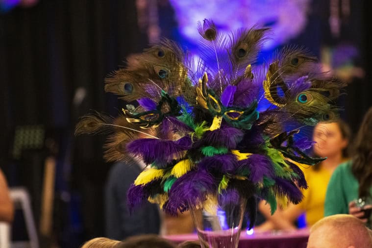 A vibrant Mardi Gras-themed decoration with colorful feathers and peacock eyes on a table. Blurred people and event setting in the background.