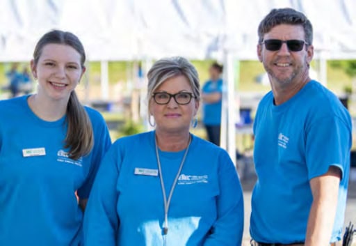 Three people in blue shirts with name tags stand together outdoors, smiling. Tables and a grassy area are visible in the background.