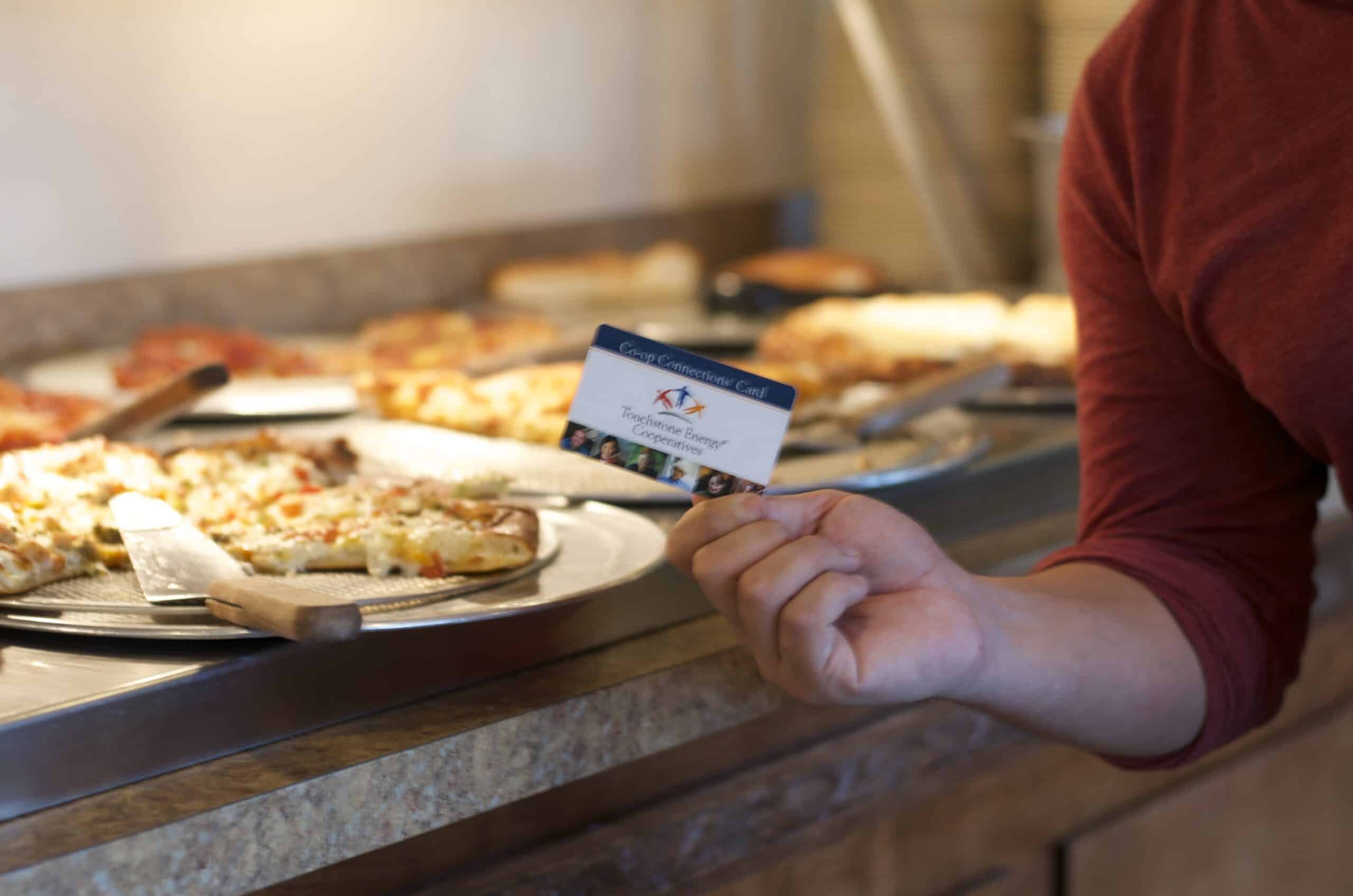 A person in a red shirt holds a Torchstone Energy Cooperatives card near a counter with several pizzas. No landmarks or historical buildings visible.