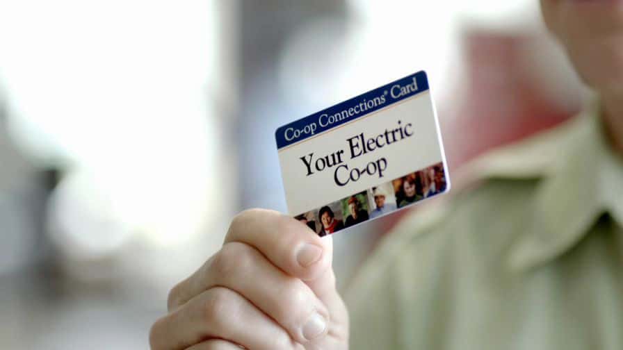 A person holds a Co-op Connections Card from "Your Electric Co-op," featuring a few faces on it and blurred background.