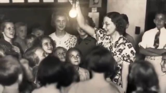 A group of children enthusiastically gather around a person holding a lit light bulb, showcasing a science lesson in a classroom setting.