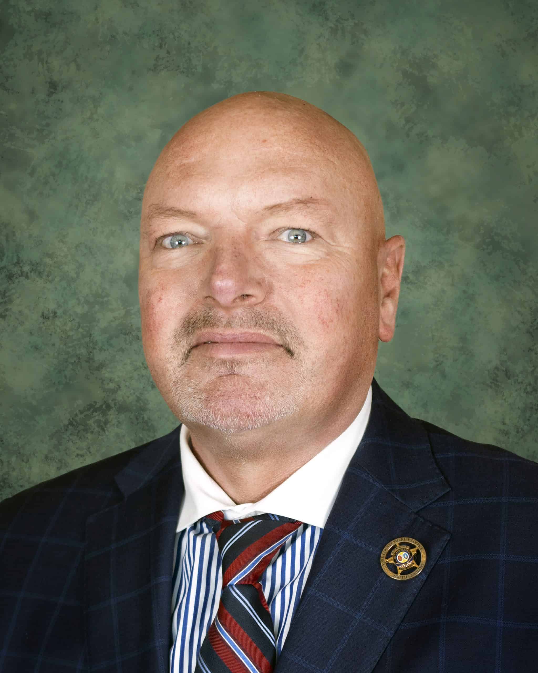 A person in a formal suit with a striped tie poses against a green background. A badge is visible on the person's lapel.