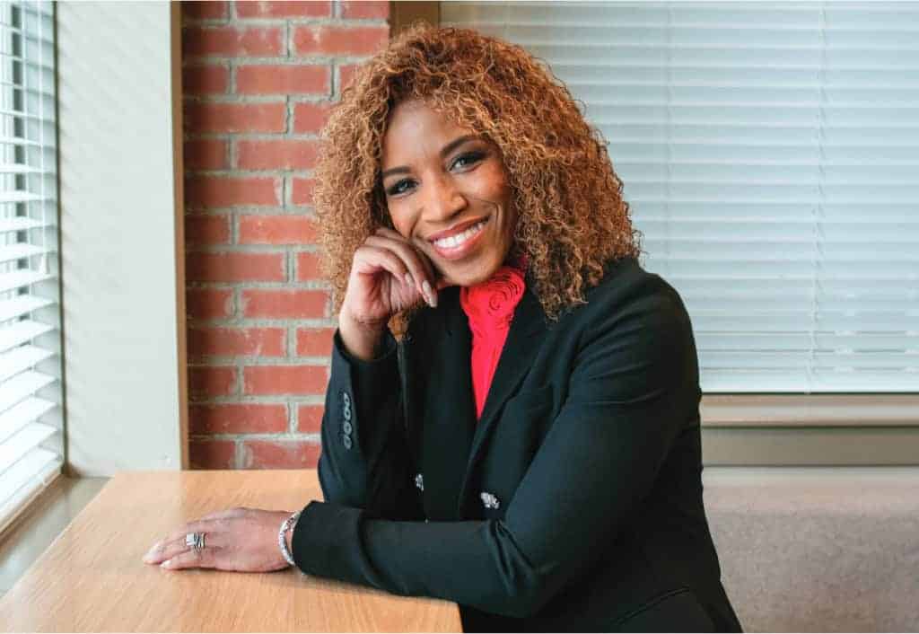 A person with curly hair smiles at a wooden table, sitting against a brick wall and window blinds. Dressed in formal attire.