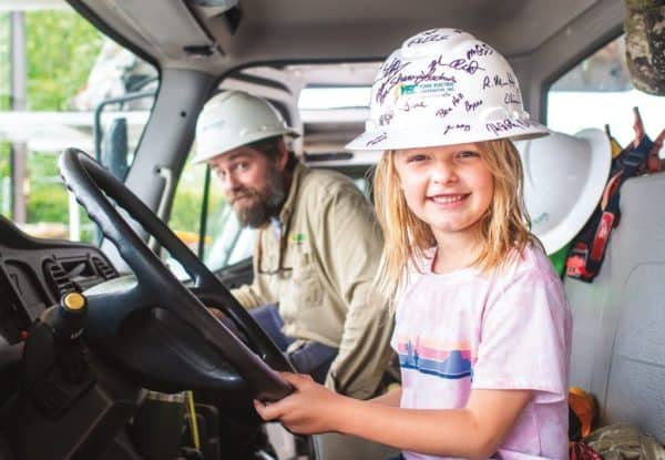 A child and a person in hard hats inside a vehicle. The smiling child is sitting at the steering wheel, posing playfully.