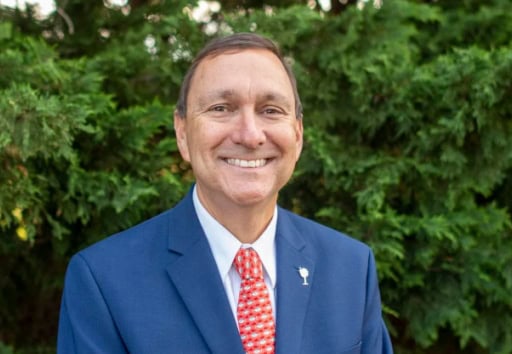 A person wearing a blue suit and red tie smiles in front of a background of green foliage. No landmarks or historical buildings are visible.