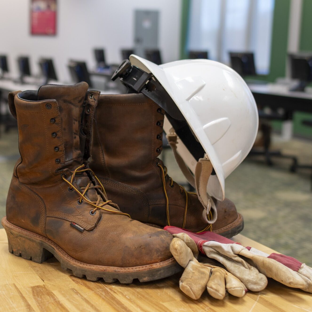 A pair of brown work boots, a white hard hat, and gloves sit on a table in a computer-equipped room.