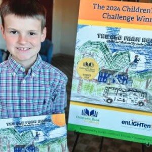 A child holding a book stands beside a poster announcing the 2024 Children's Book Challenge Winner, featuring the same book's cover artwork.