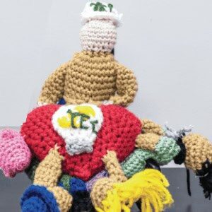 The image depicts a crocheted scene with colorful figures, including a person wearing a white hat and beige clothing, kneeling around a red and white centerpiece.