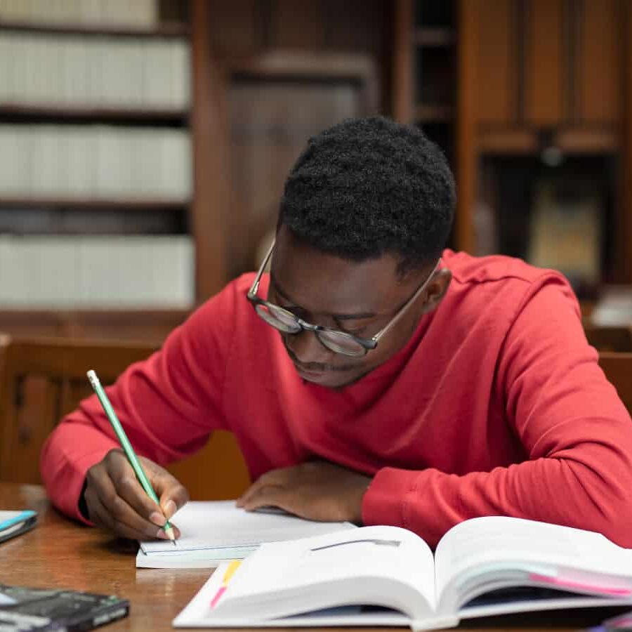 A person in a red shirt studies at a wooden table in a library filled with bookshelves and various materials.