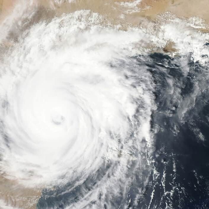 A massive cyclone swirls over the ocean, nearing a desert coastline. Dense clouds indicate intense weather conditions approaching. No landmarks visible.