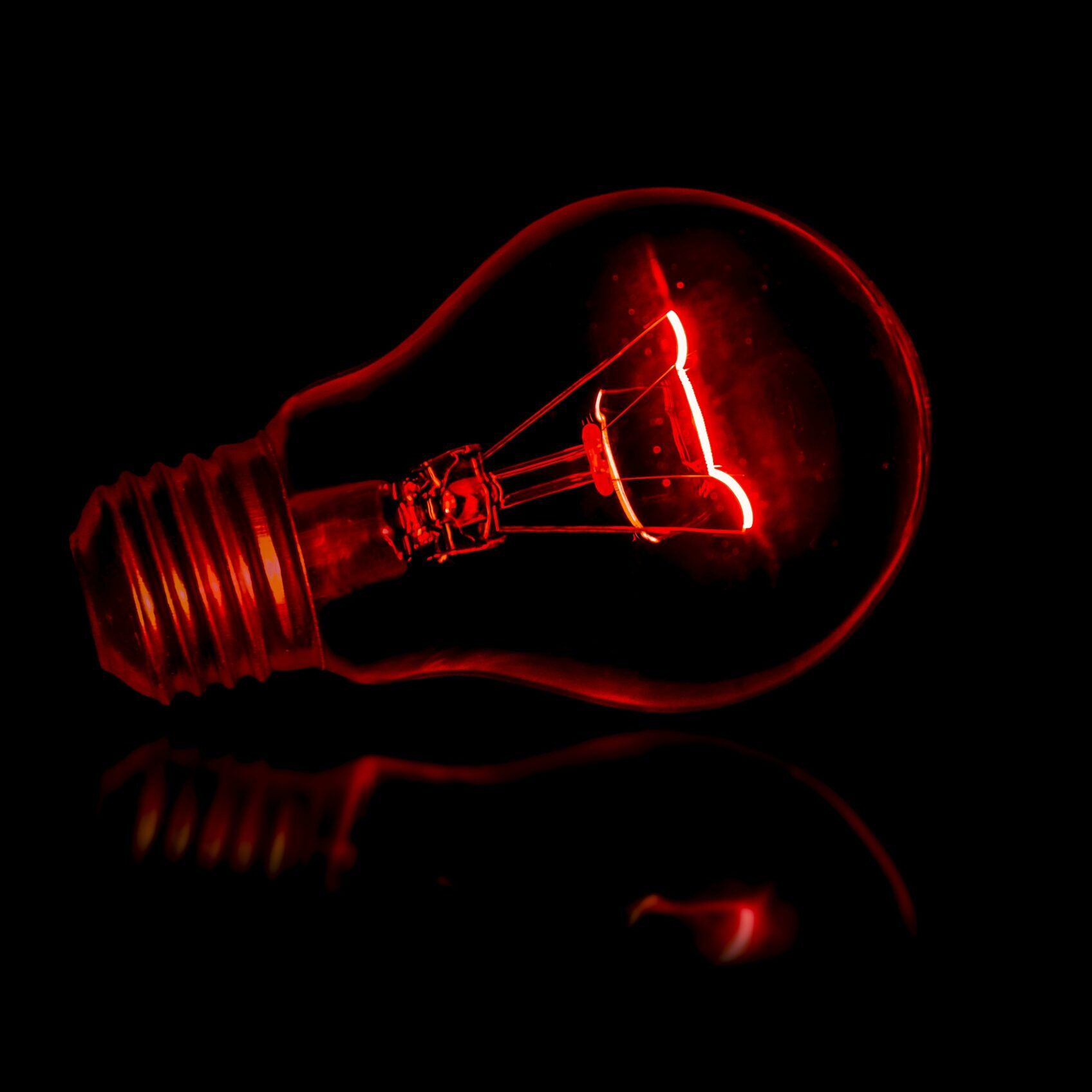 A red incandescent light bulb glows brightly against a dark, reflective background, highlighting the intricate filament inside.