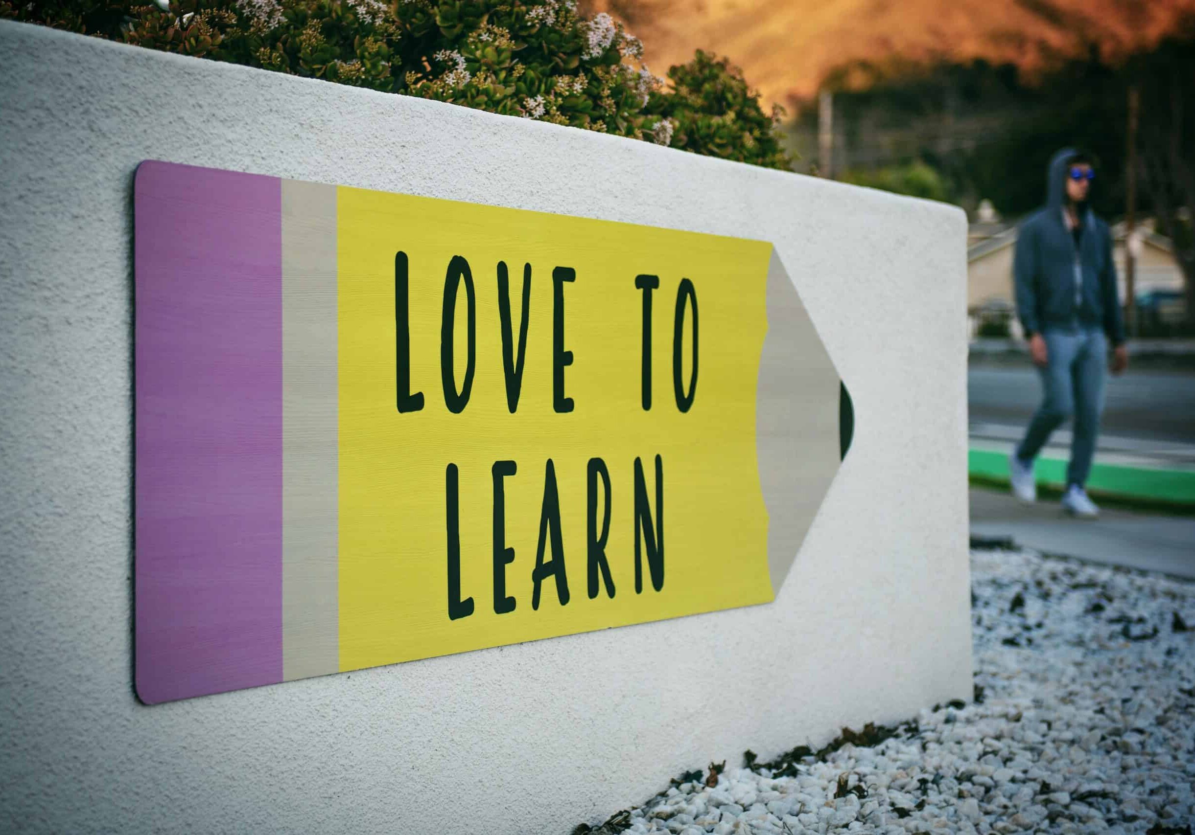 A sign shaped like a pencil with "Love to Learn" in bold letters. A person walks in the background on a sidewalk.