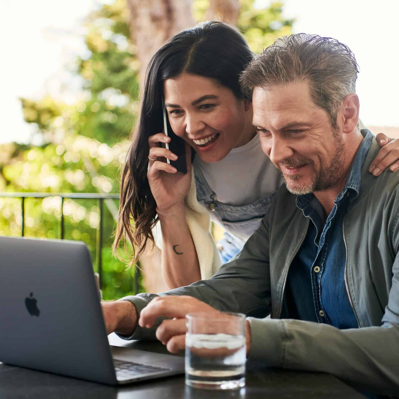 Two people smiling at a laptop screen outdoors, one on the phone. Both appear engaged and happy. Glass of water on table nearby.