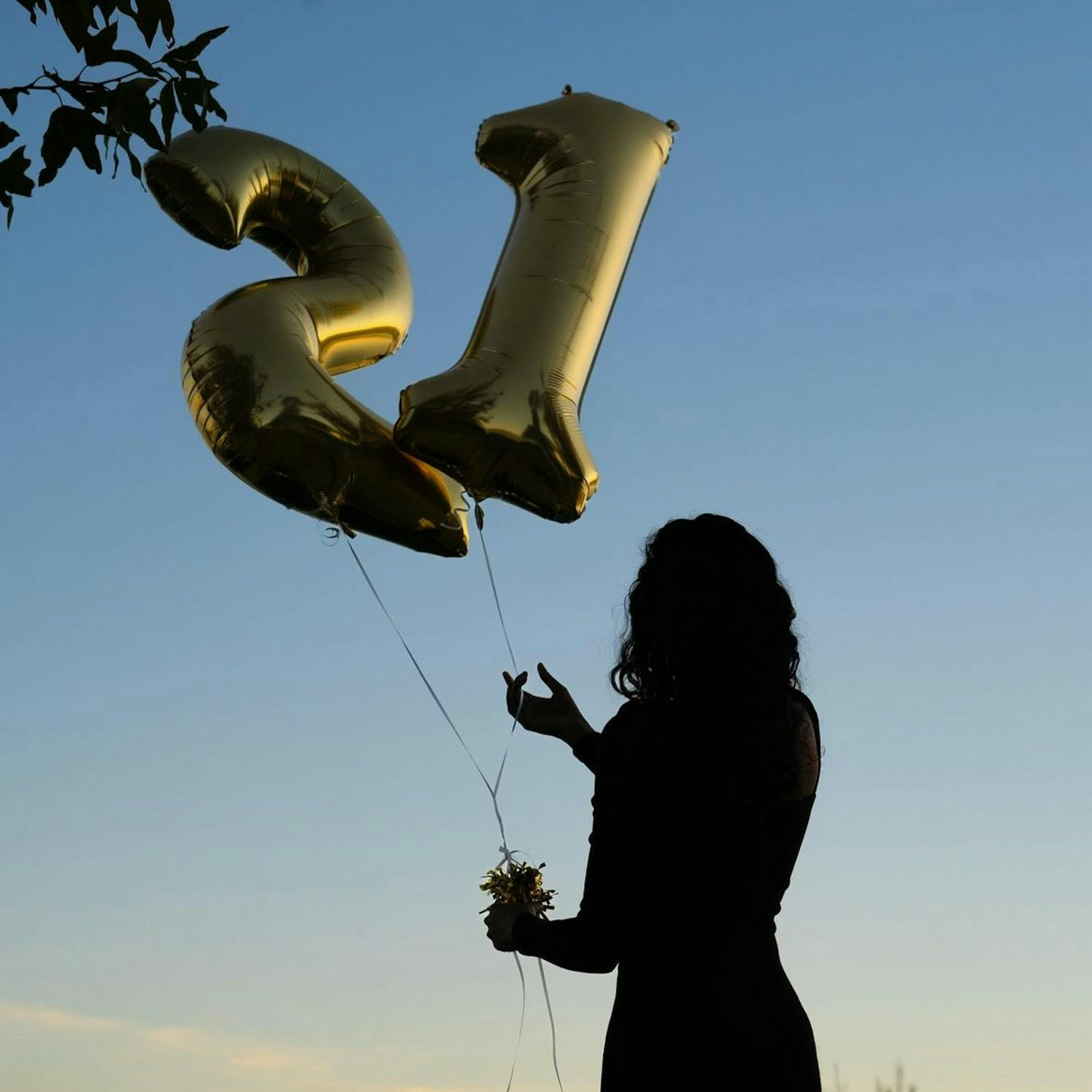 A person holding golden "21" balloons stands against a clear blue sky, with leaves visible in the top corner. No landmarks are present.