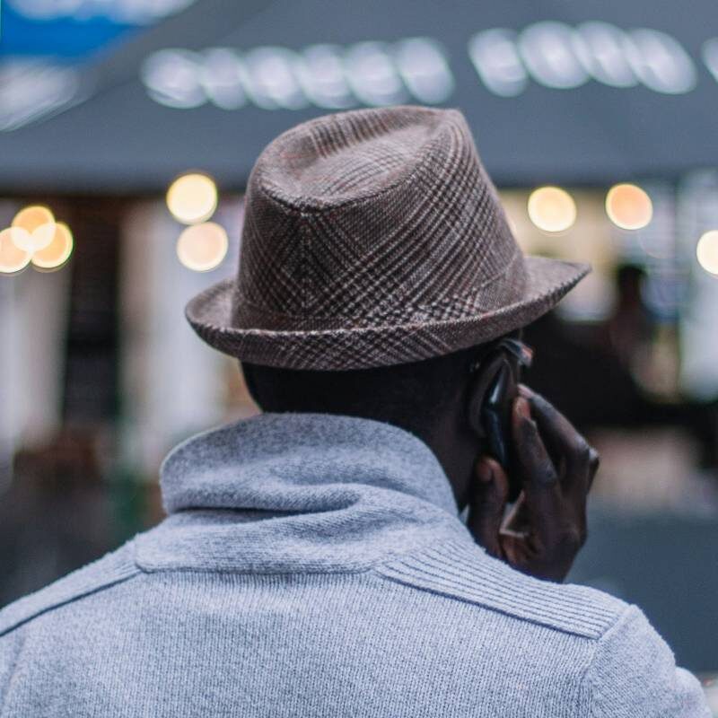 A person in a gray sweater and hat talks on a phone, standing near a street food market with blurred lights in the background.