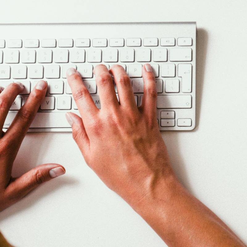 A person is typing on a white computer keyboard, with both hands visible on the keys against a plain background.