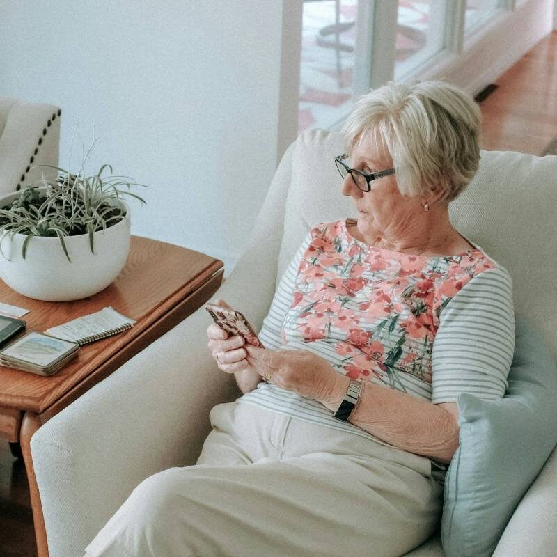 A person with short hair and glasses sits on a couch, looking at a phone. A plant and notepad are nearby.