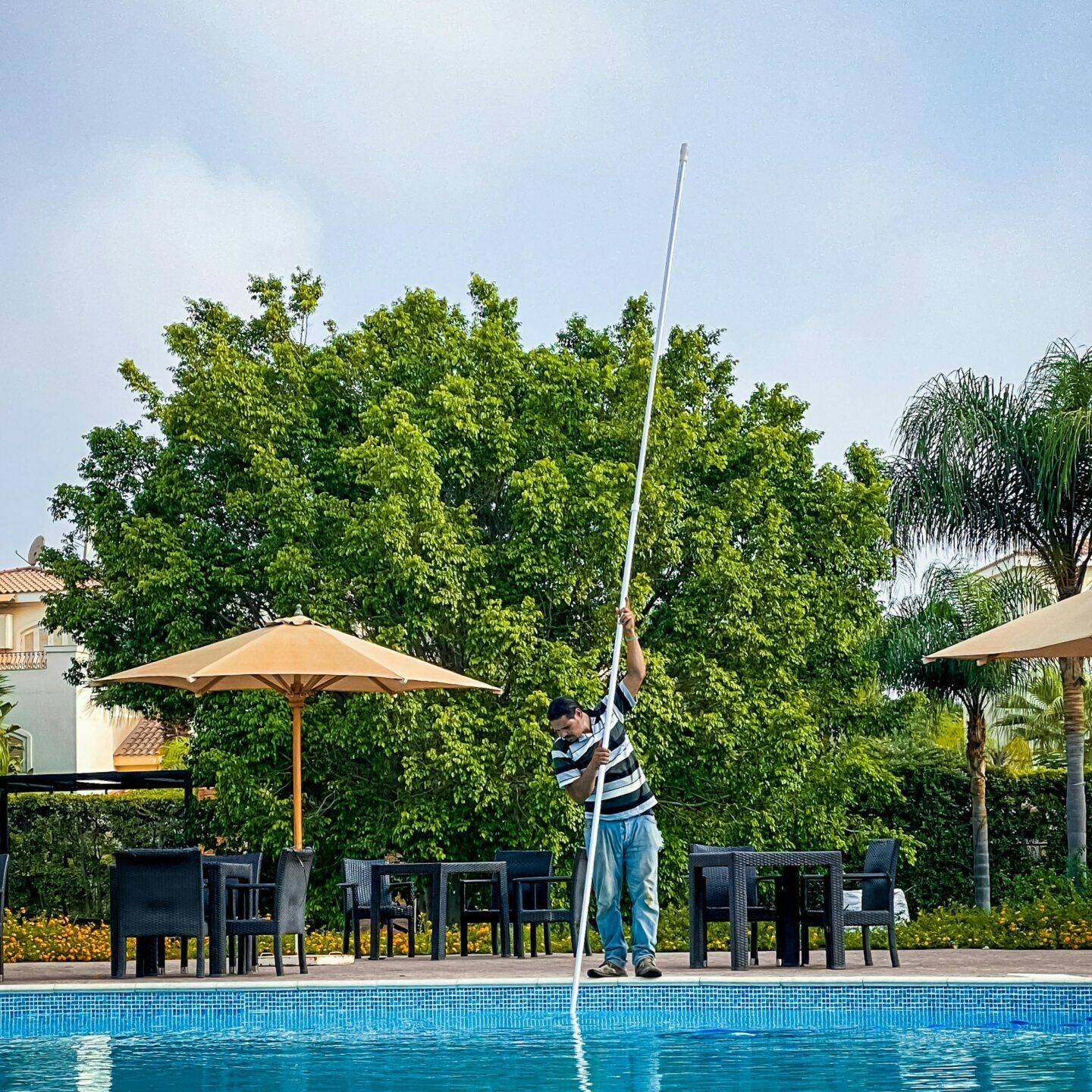 A person cleans a swimming pool with a long pole. There are chairs and umbrellas beside the pool, and green trees in the background.