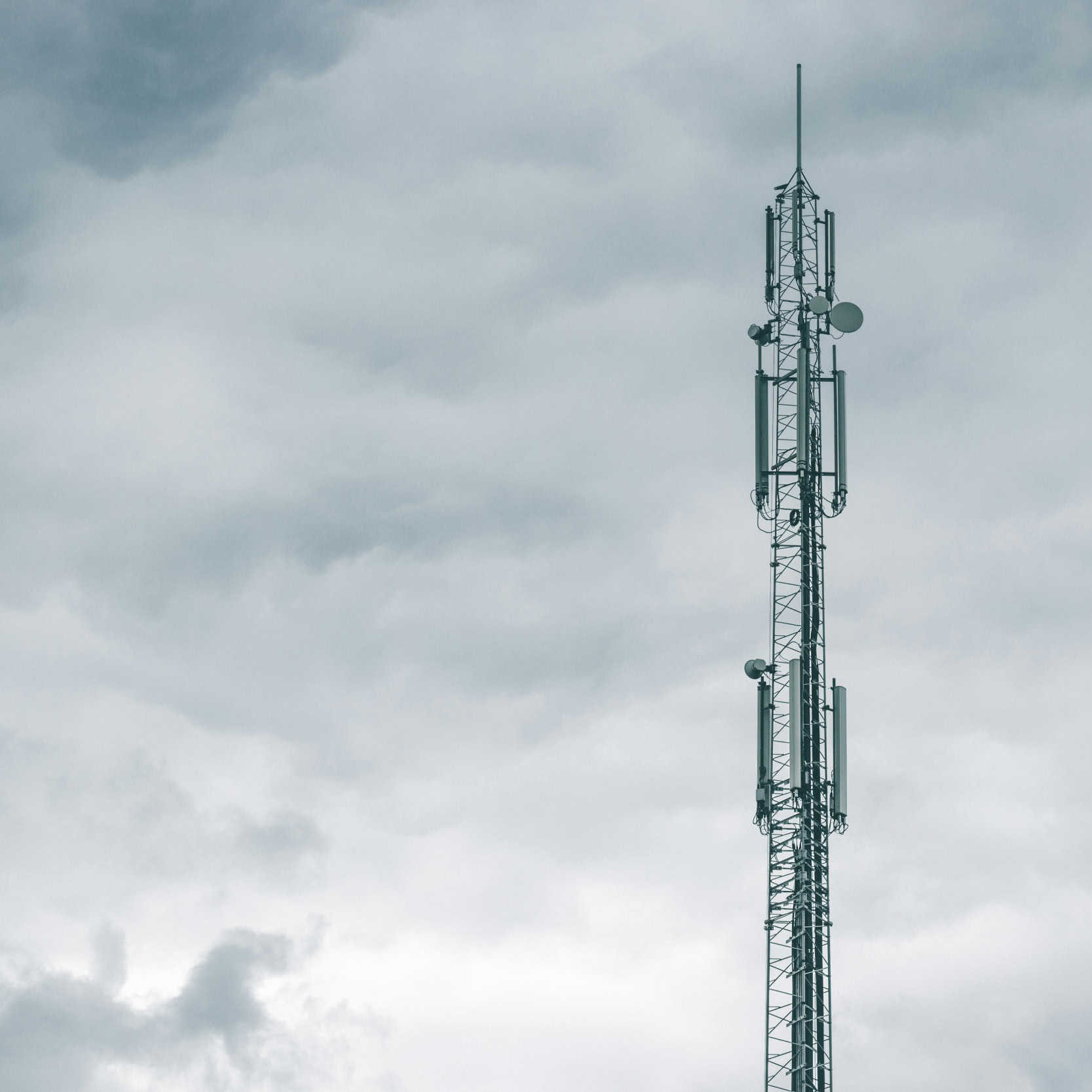 A tall telecommunications tower stands against a backdrop of overcast, cloudy sky. The structure appears isolated, with various antennas attached.