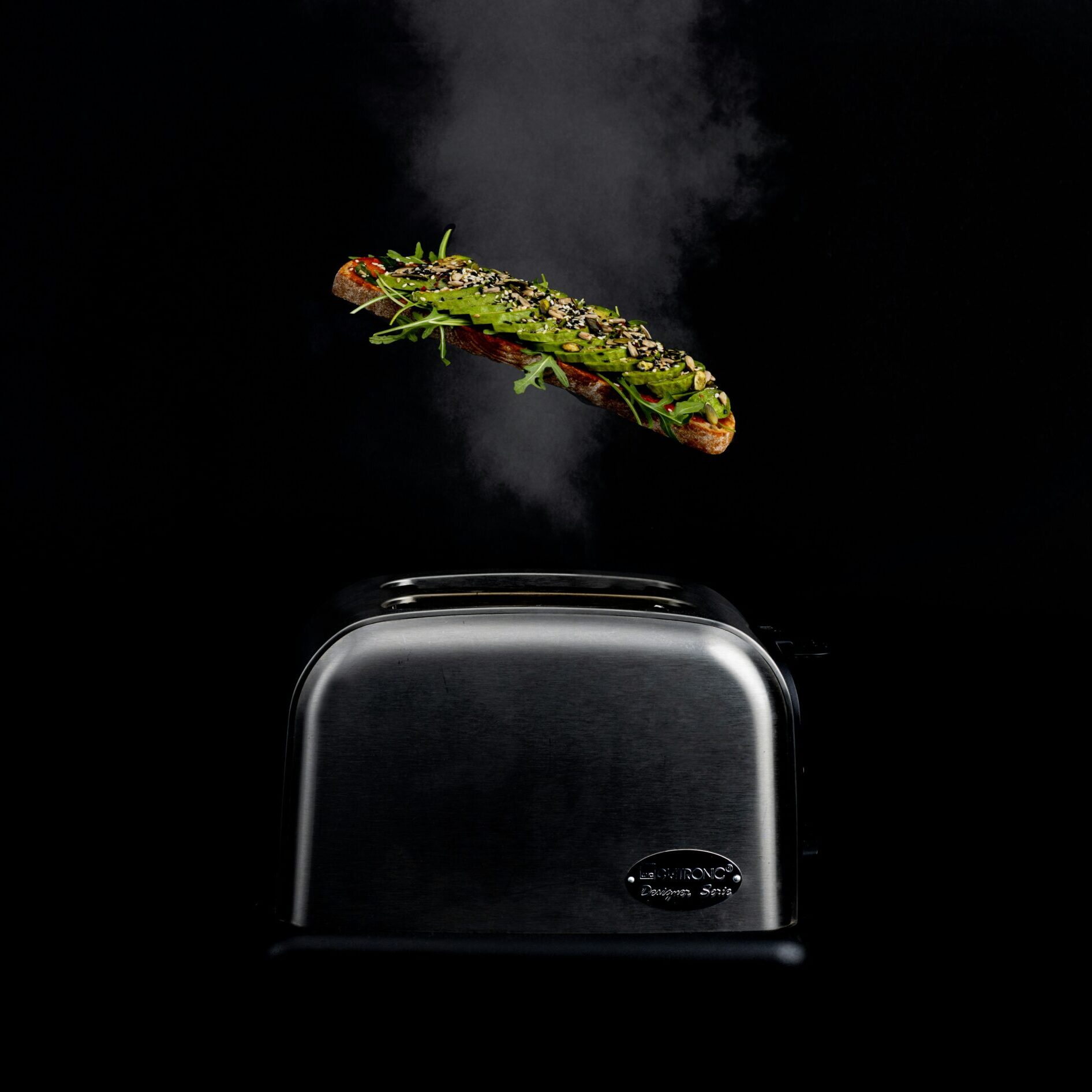 A slice of toast with green toppings levitates above a stainless steel toaster, against a dark background with visible steam.