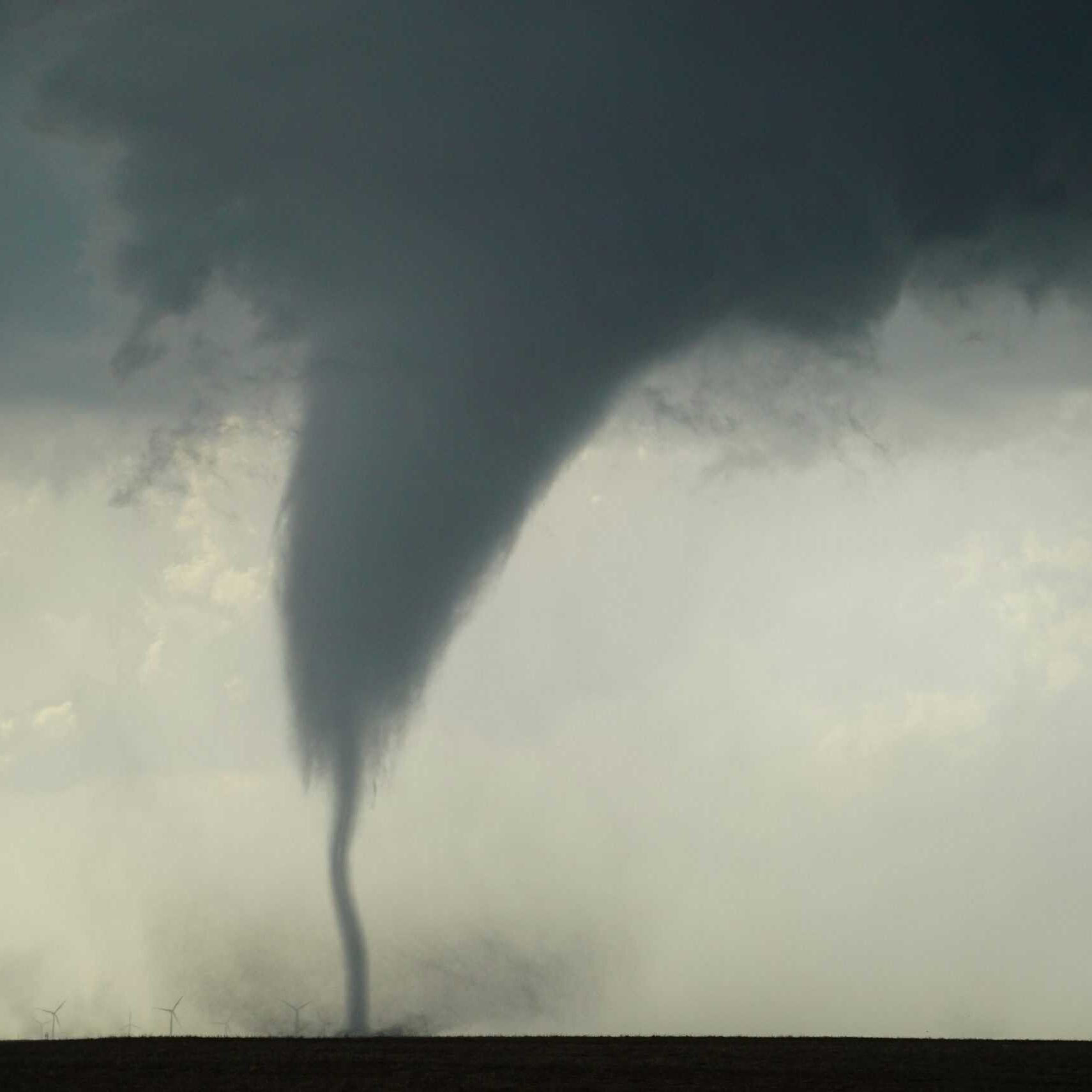 A large tornado is descending from a dark stormy sky, touching the ground in an open field with wind turbines in the distance.