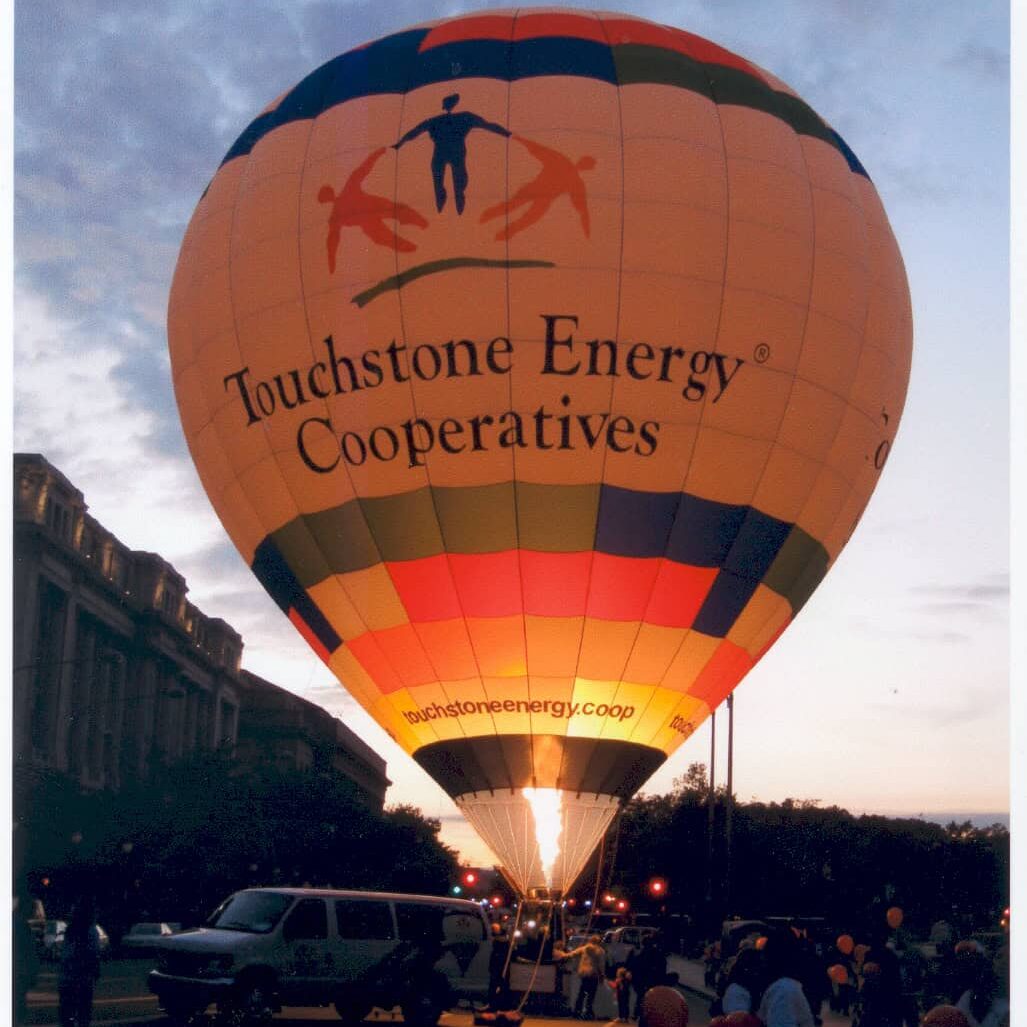 A hot air balloon with "Touchstone Energy Cooperatives" logo, lit at dusk, surrounded by people. Nearby vehicles and a historical building are visible.