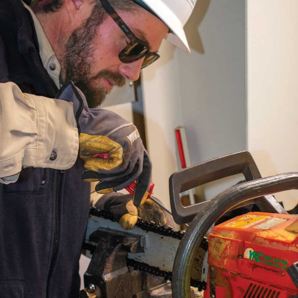 A person wearing sunglasses and a hard hat sharpens a chainsaw chain using a file. They are focused on their task.