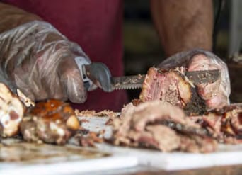 A person wearing gloves slices cooked meat with a knife on a white cutting board, focusing on a close-up view of the preparation process.