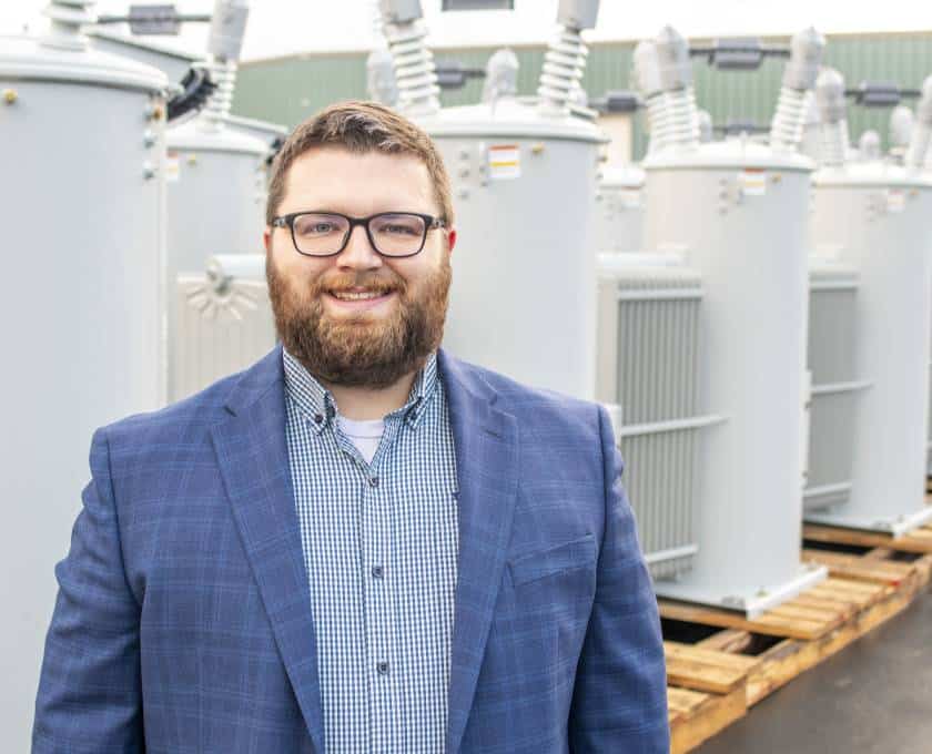 A person with glasses and a beard stands smiling near large industrial equipment outdoors, wearing a blue suit jacket.