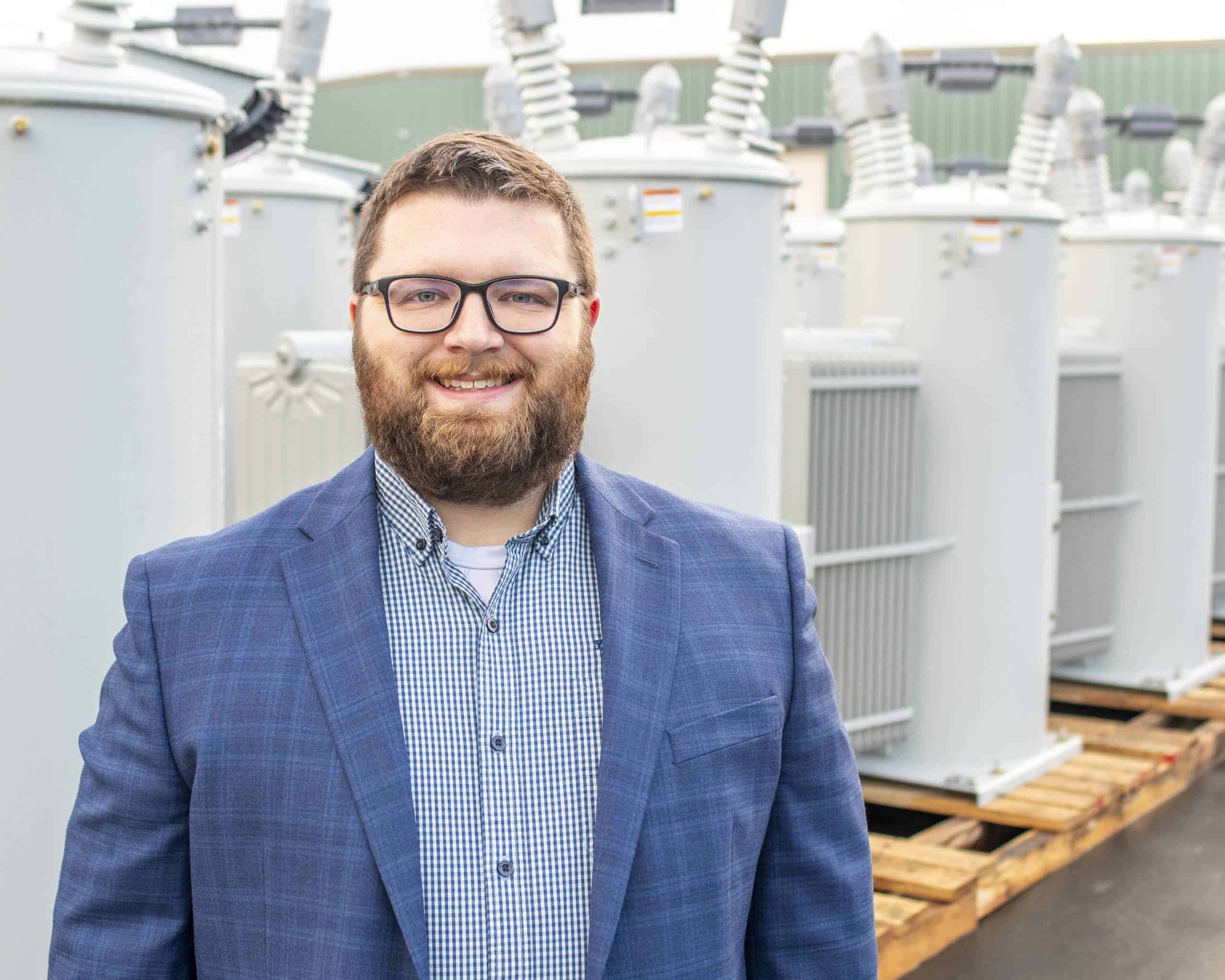 A person in a suit stands outdoors, smiling, with a background of industrial equipment on wooden pallets.