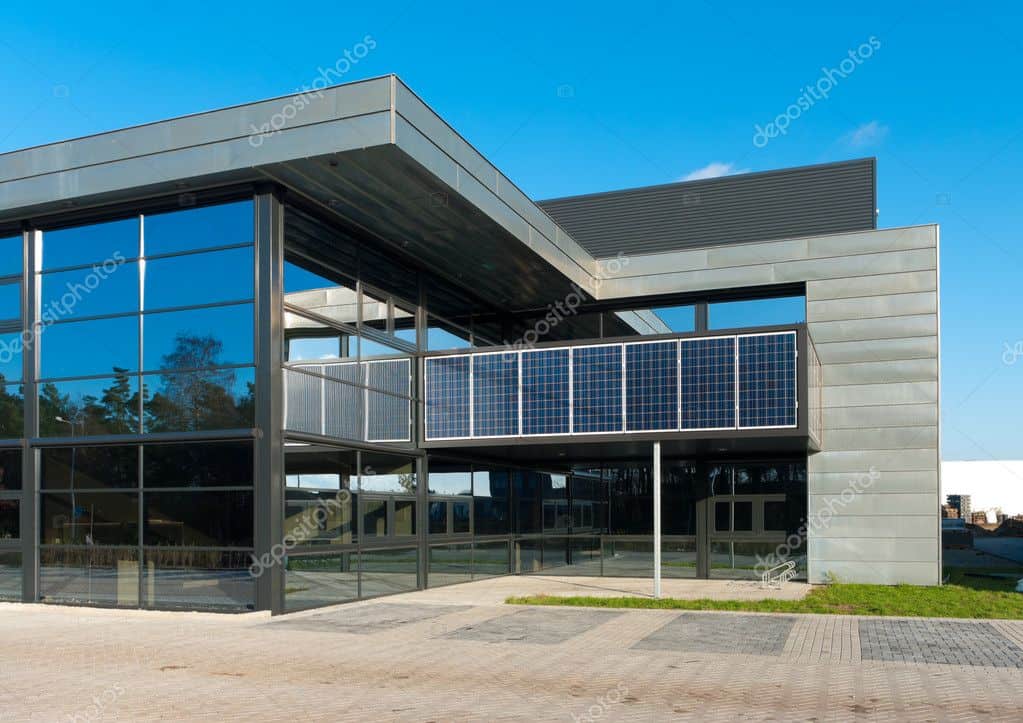 A modern building with large glass windows and solar panels on its facade, set against a clear blue sky and surrounded by greenery.