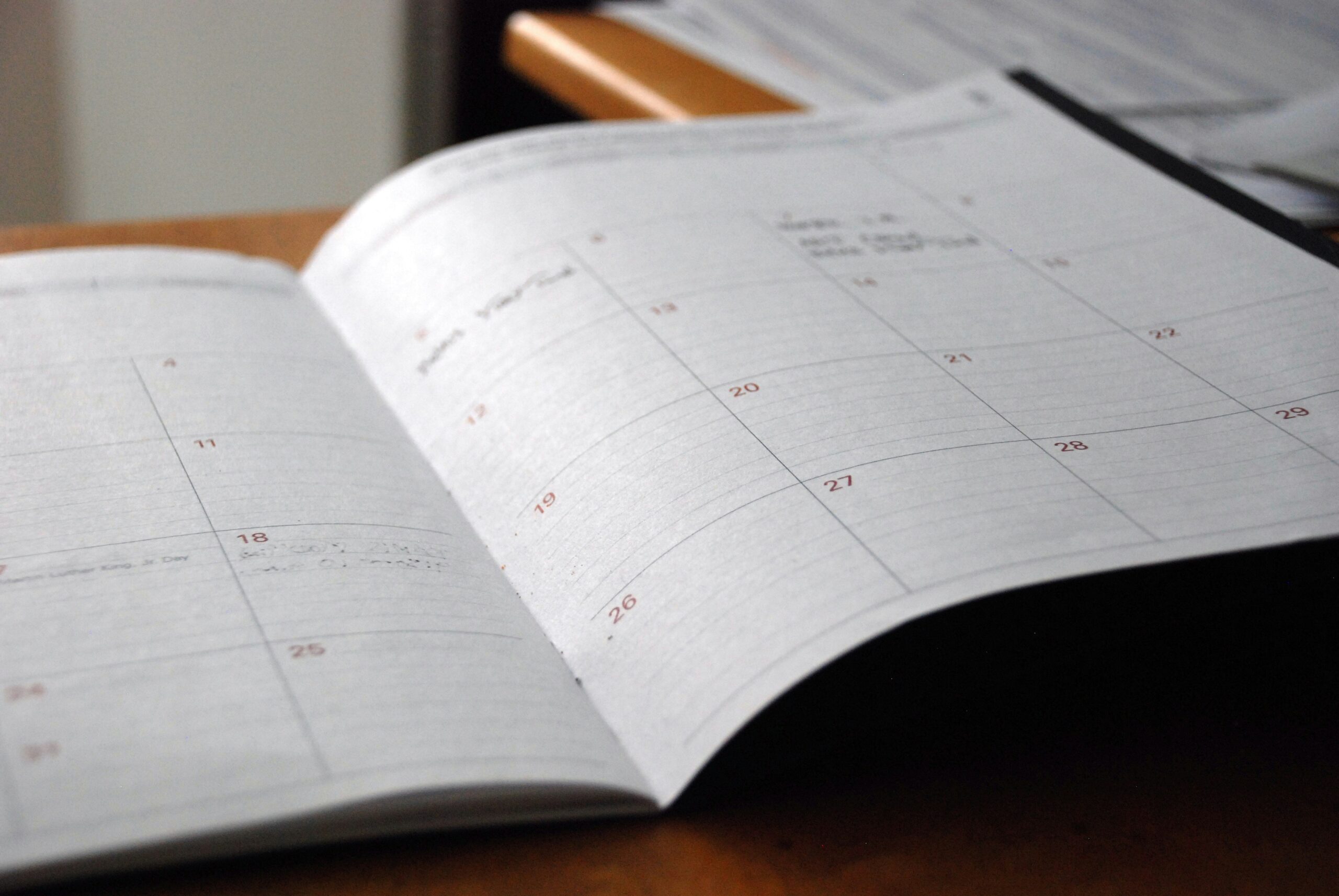 An open planner lies on a wooden table, displaying a weekly view with dates written in red, set in a softly lit room.