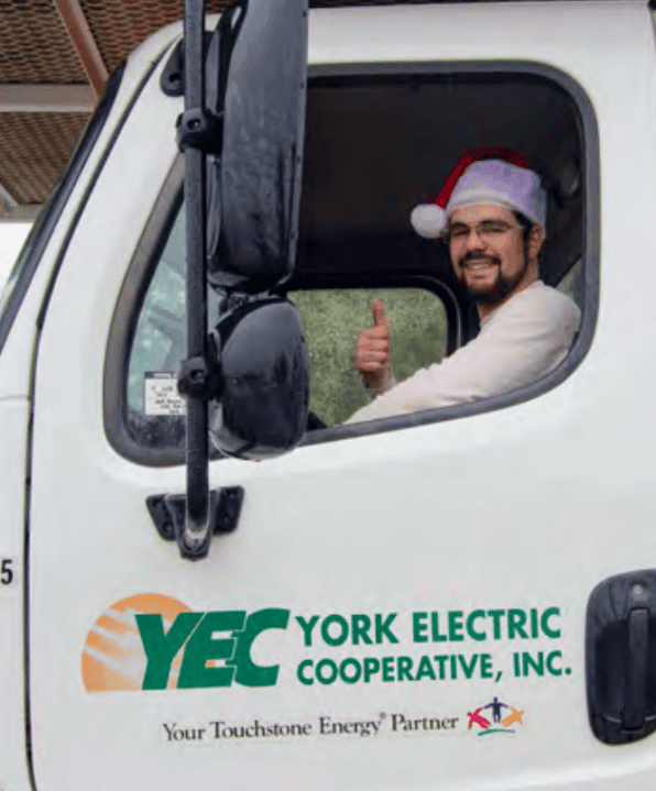 A person wearing a Santa hat gives a thumbs-up inside a York Electric Cooperative truck cab, with the company's logo visible on the door.