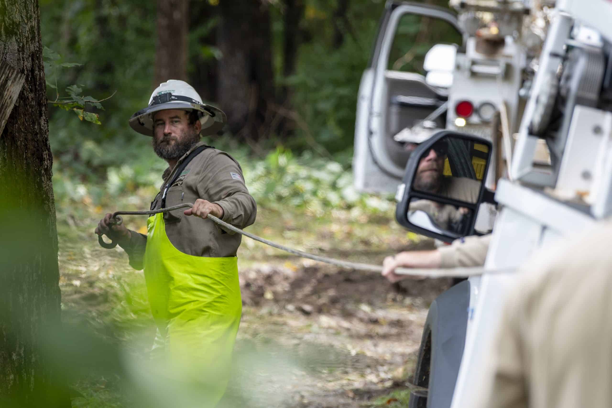 A person in a hat and bright overalls pulls a rope near a truck in a wooded area. Another person assists from the vehicle.