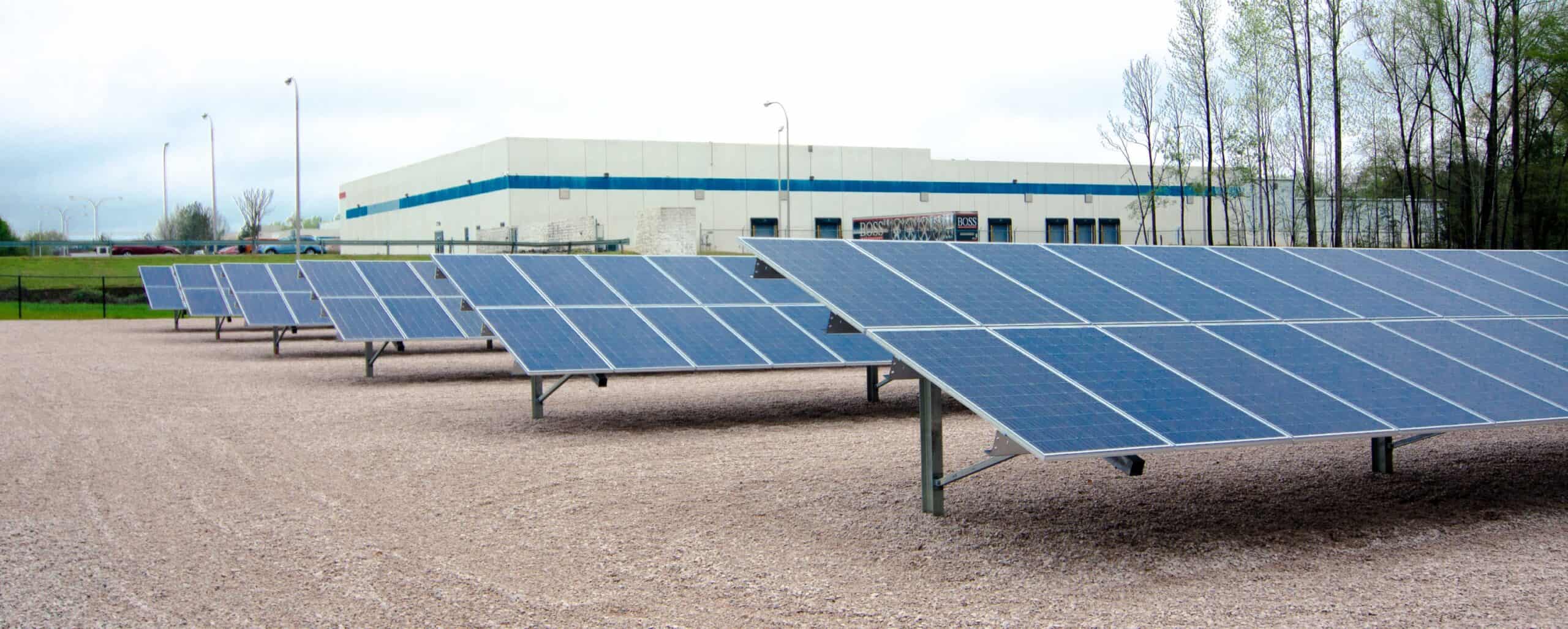 A row of solar panels on gravel with trees and a large building in the background, under a cloudy sky.