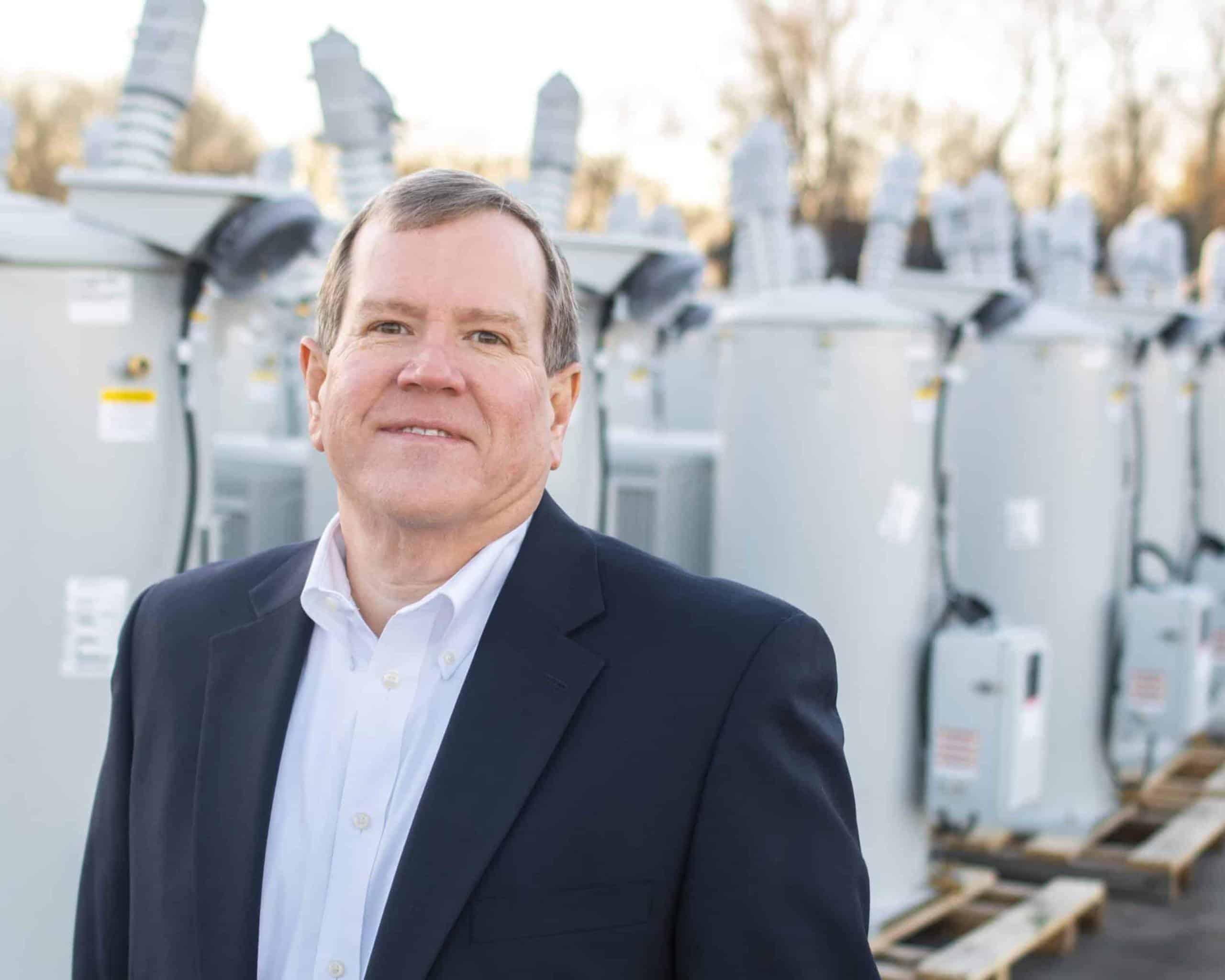 A person in formal attire stands near a row of industrial transformers, with a blurred natural background, during daytime.