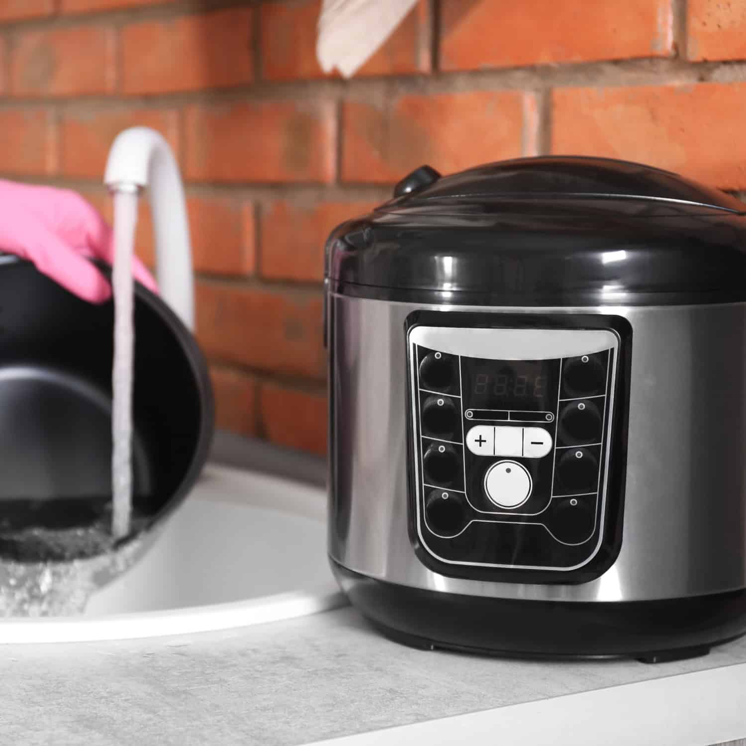 A person wearing pink gloves rinses a pot at a sink near a countertop multicooker, with a brick wall backdrop.