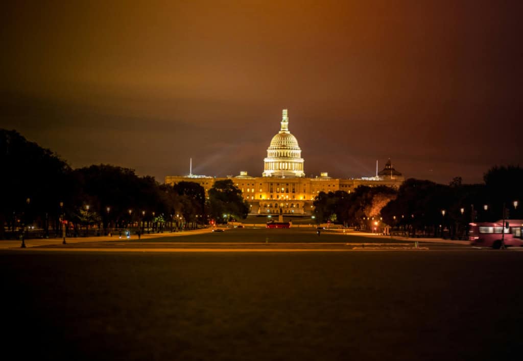 The image shows the U.S. Capitol at night, illuminated with a warm glow, surrounded by trees and a dark sky.