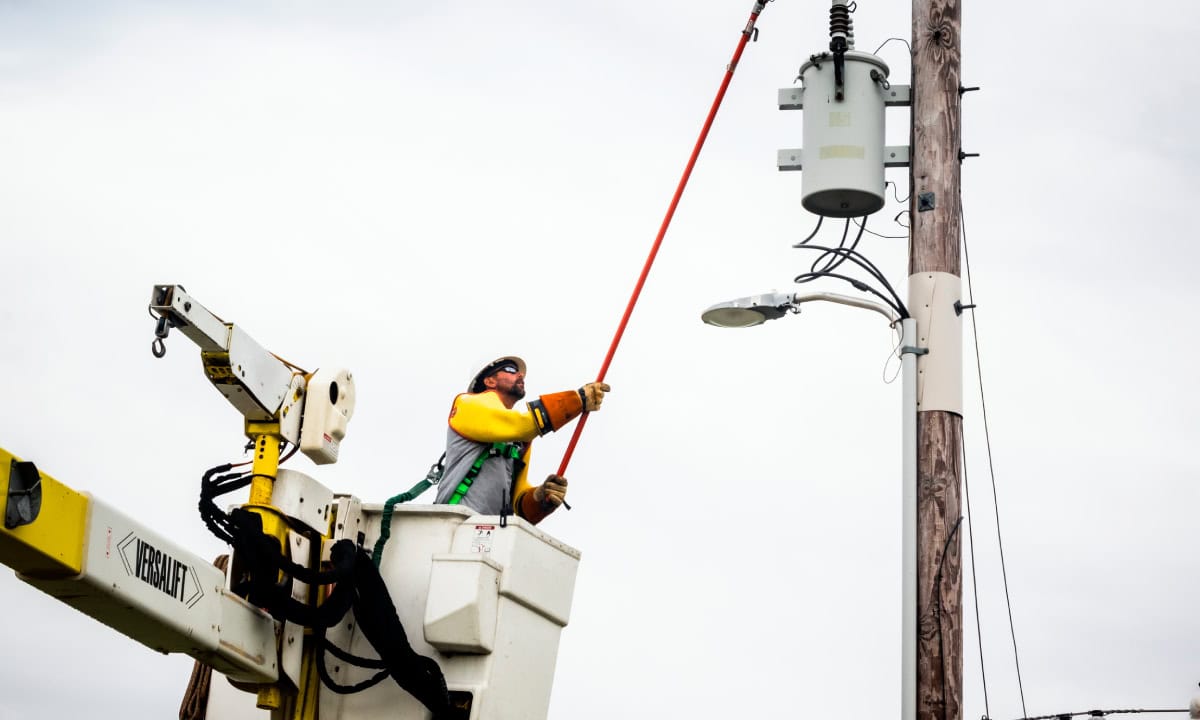 A person in a lift uses a long pole to work on a utility pole's electrical equipment against a cloudy sky.