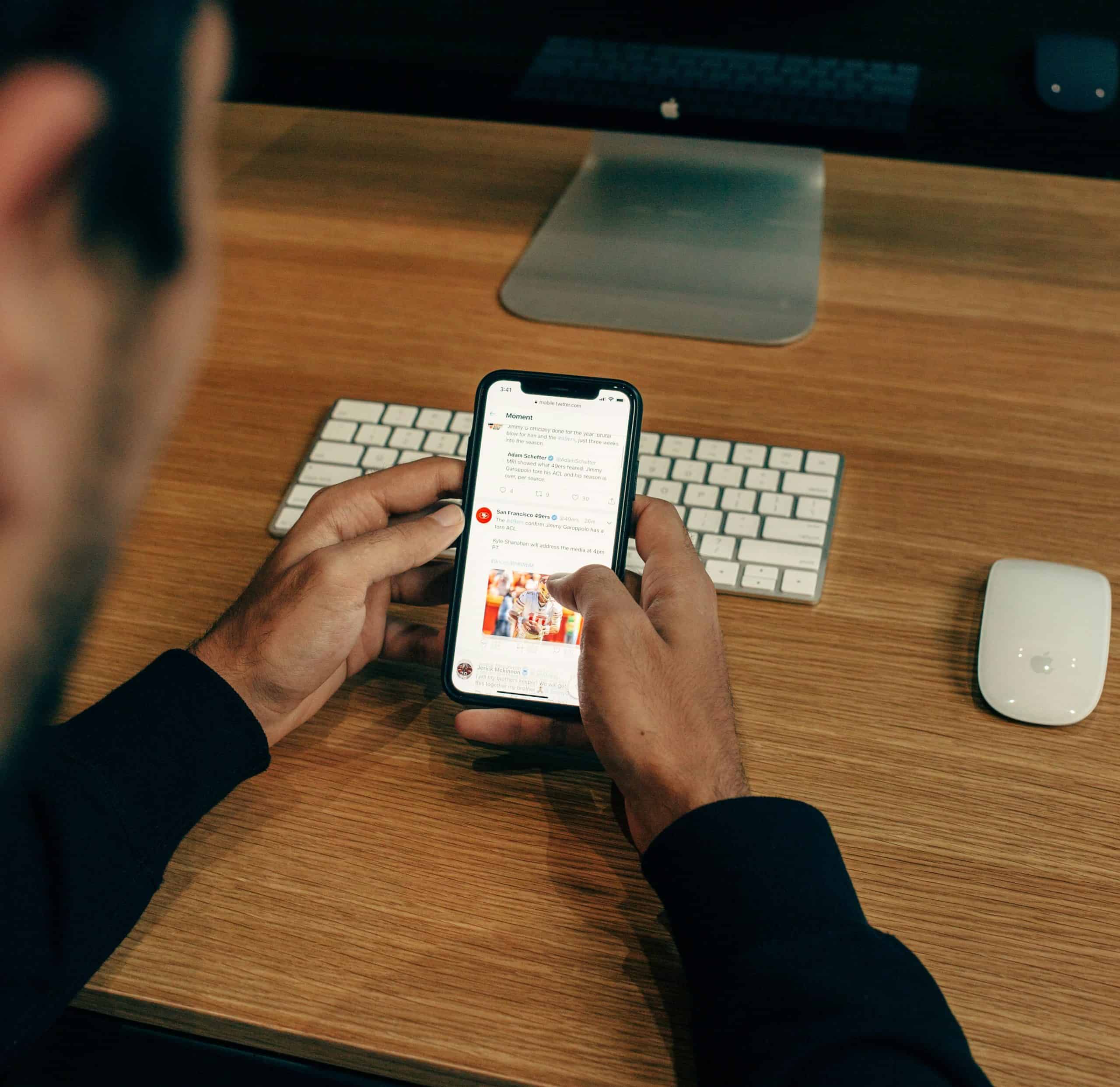 A person using a smartphone while seated at a wooden desk with a keyboard, mouse, and monitor in the background.