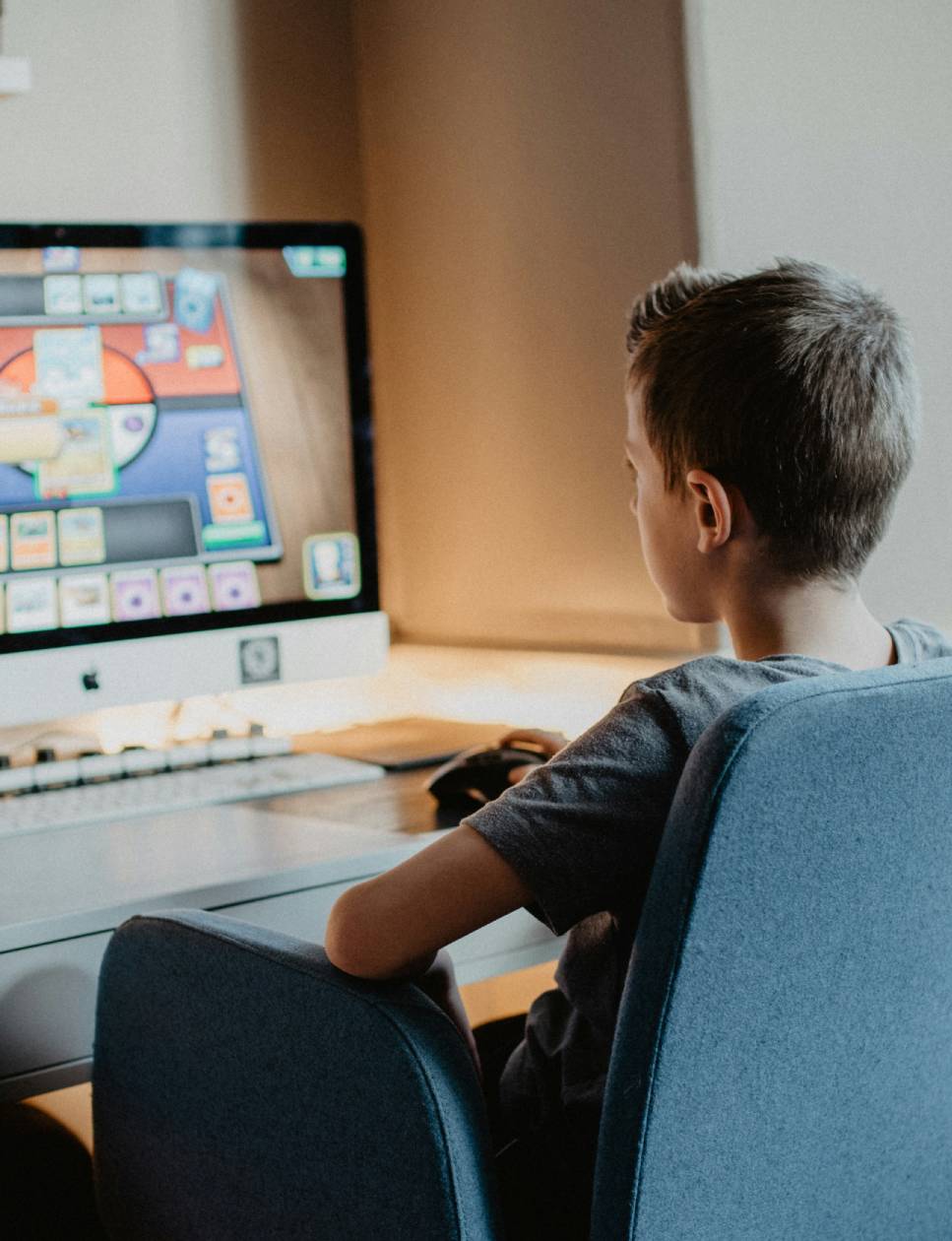 A child sits in front of a computer, absorbed in an online card game, with a mouse and sleek gray chair visible.