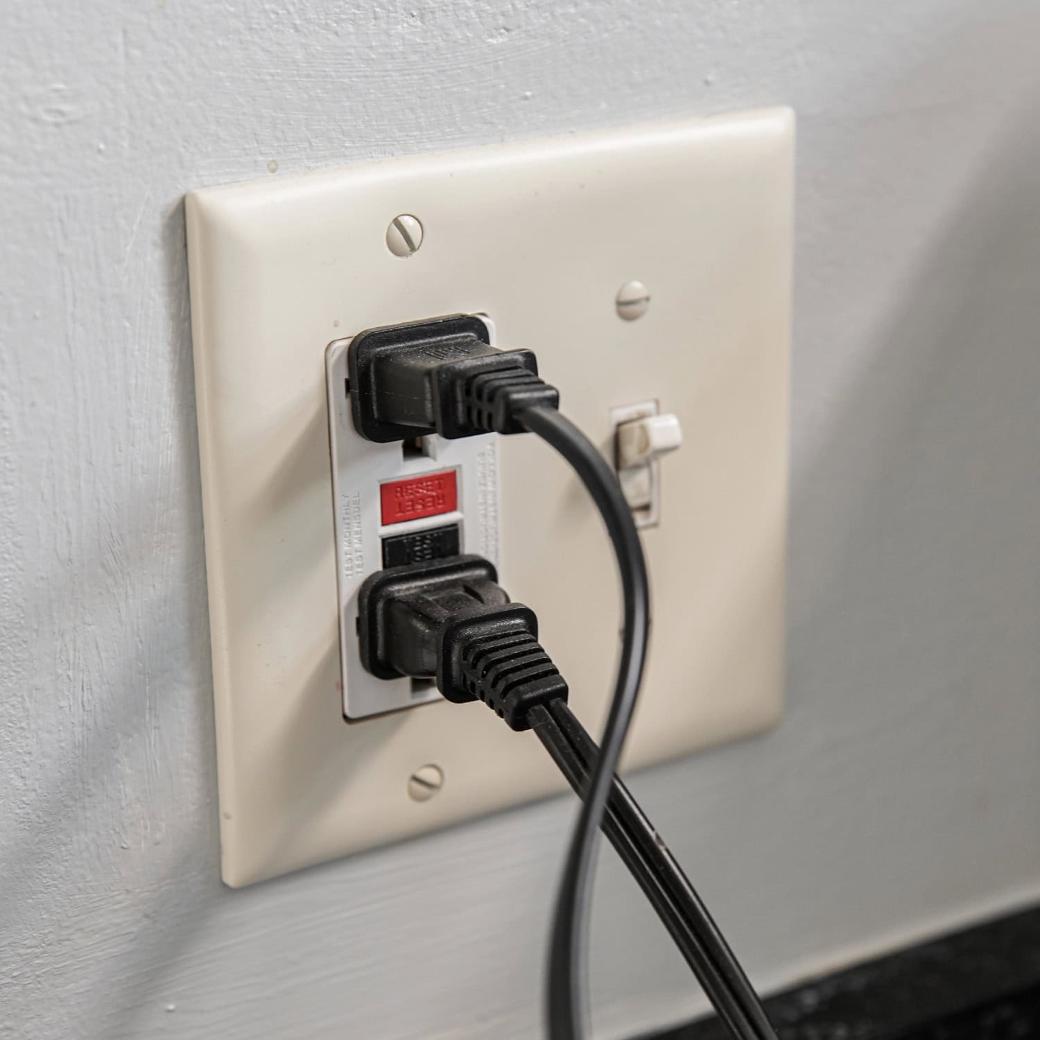 A wall outlet with GFCI has two black plugs inserted. The outlet includes test and reset buttons on a light switch plate.