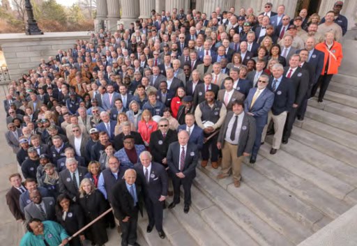 A large group of people gathers on the steps of a historical building with columns, possibly a government or institutional setting.