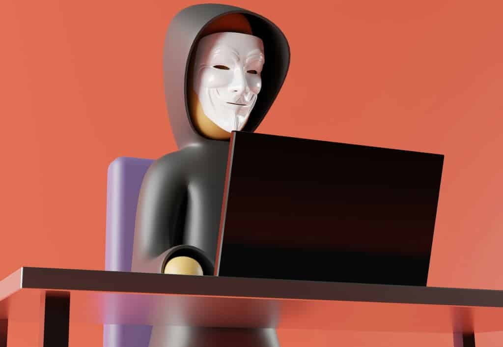 A person with a white mask and hood uses a laptop at a table in front of a solid orange backdrop.