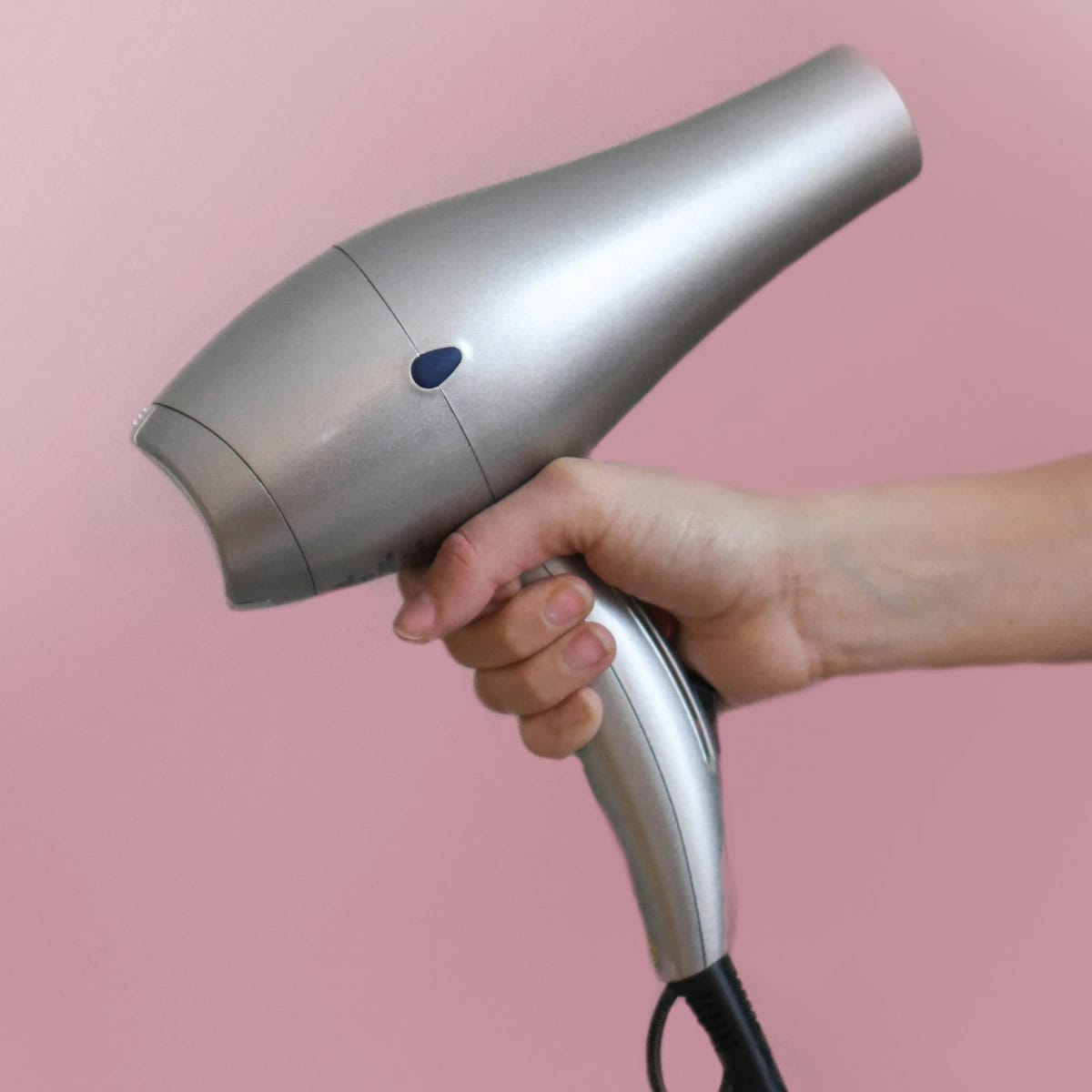 A person holding a silver hairdryer against a pink background, with a simple design and a visible power button.