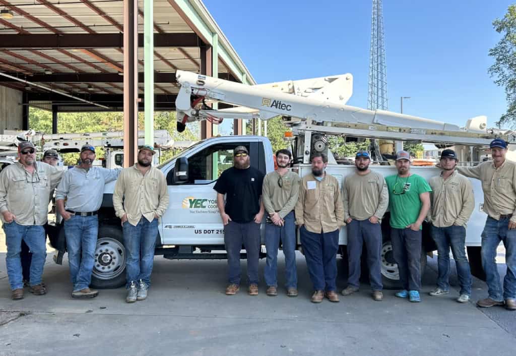 A group of ten people stand in front of a utility truck with "YEC" branding, under a metal structure. A tower is visible.