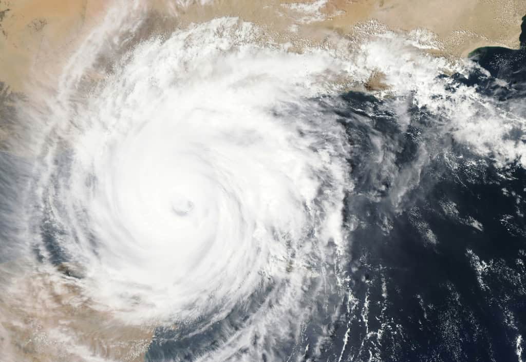 A massive cyclone swirls over the ocean, nearing a desert coastline. Dense clouds indicate intense weather conditions approaching. No landmarks visible.